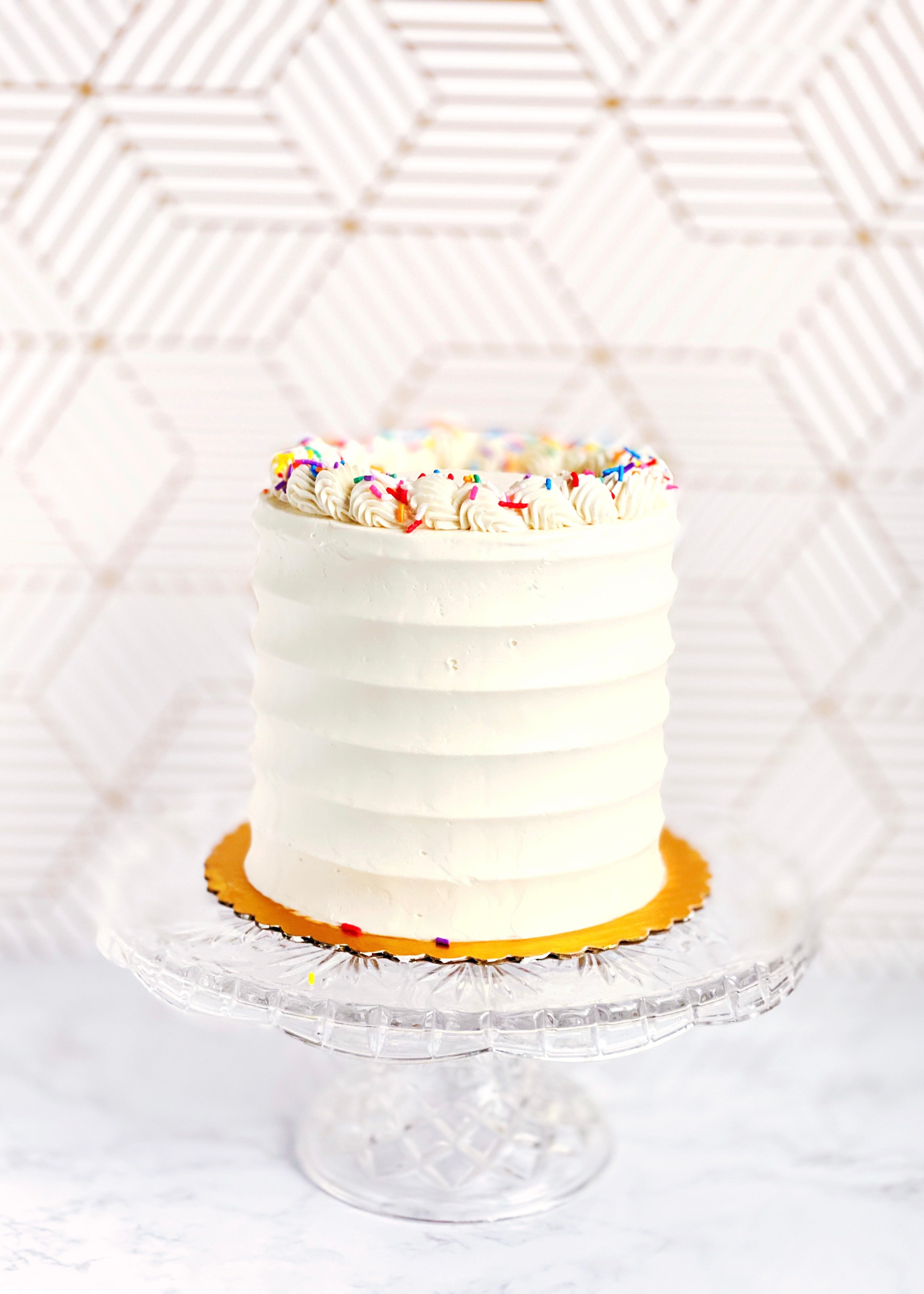 White cake with colorful sprinkles on a clear cake stand against a white geometric patterned background