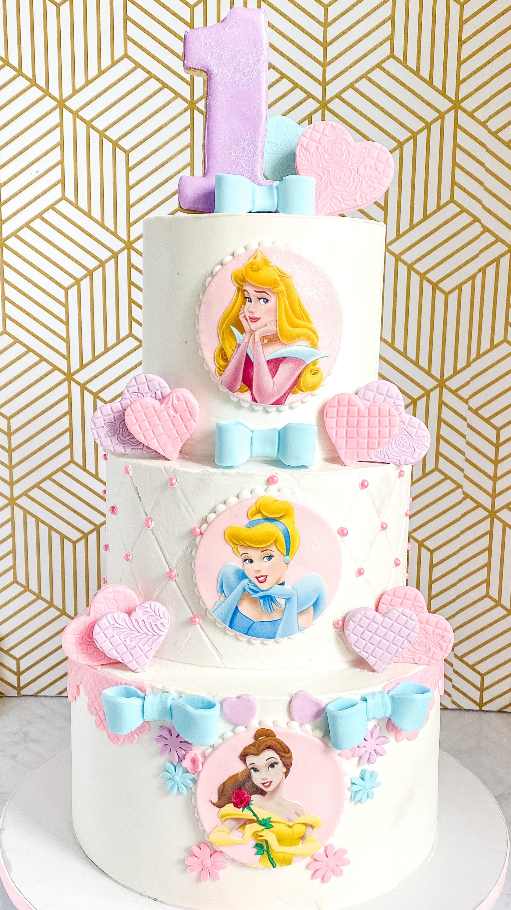 Three-tiered birthday cake with princess images against a gold geometric patterned wall.